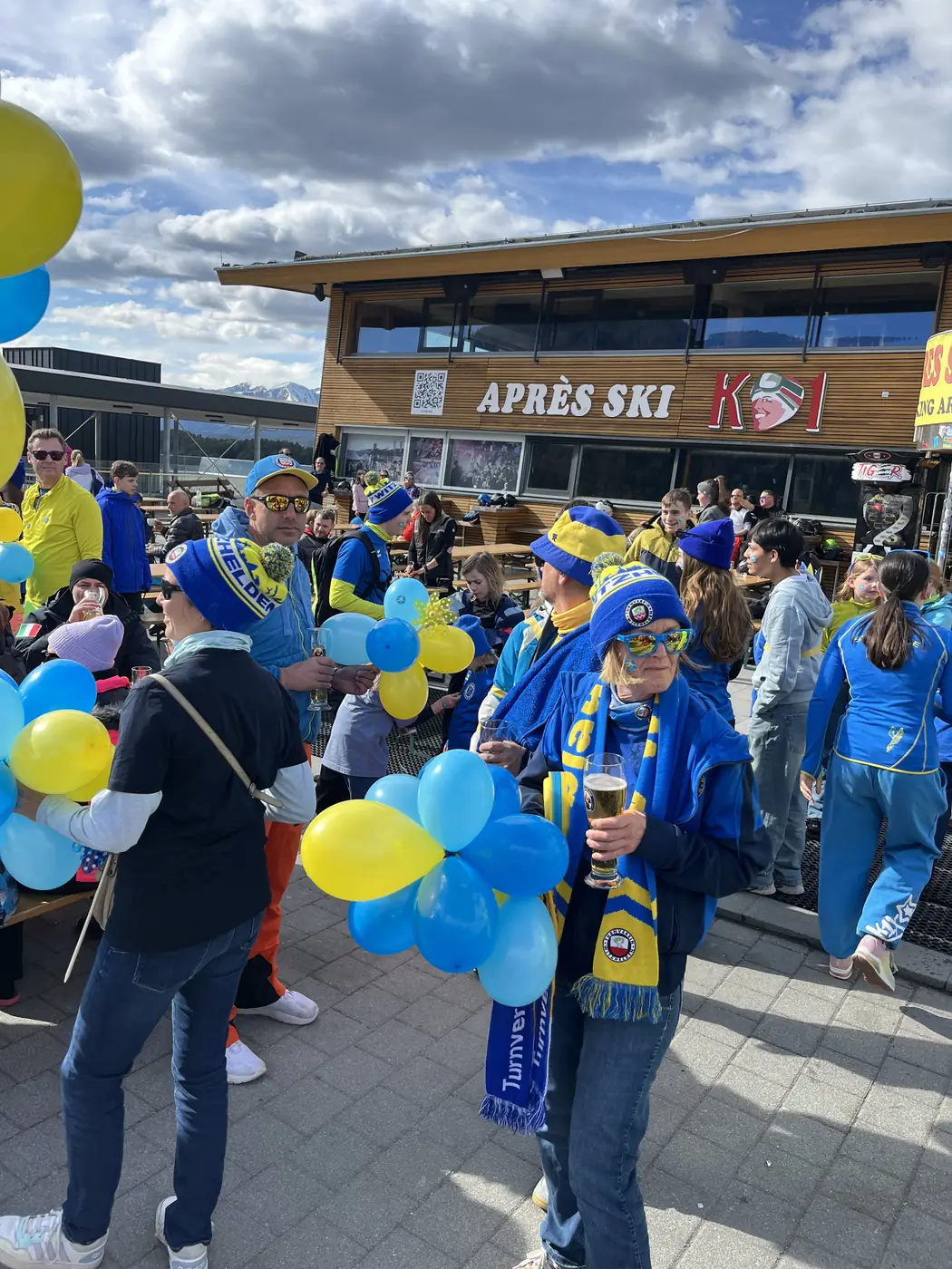 Menschen mit blauen und gelben Luftballons vor der Après-Ski-Hütte K1