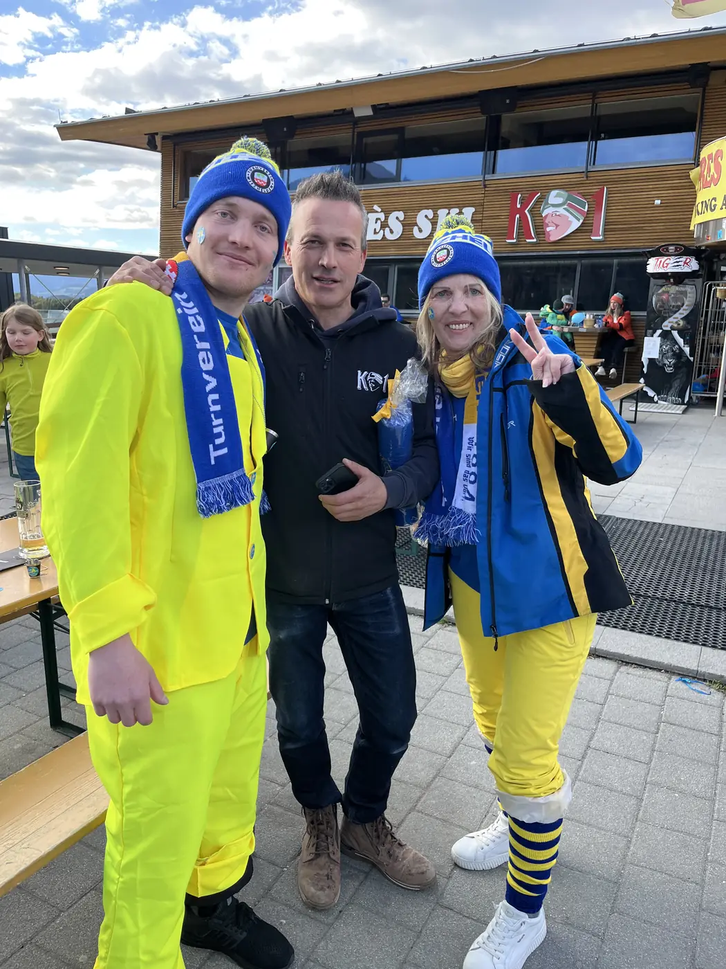 Drei Personen in Vereinskleidung bei Sonnenschein vor der Skipiste