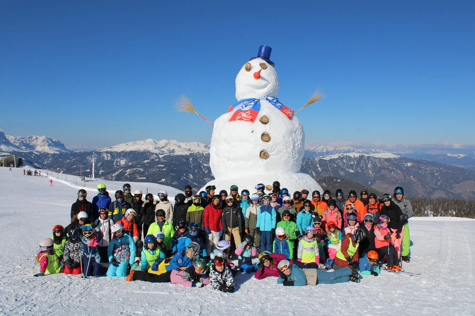 Grosse Gruppe von Skifahrerinnen und Skifahrern aller Altersgruppen vor einem riesigen Schneemann auf dem Kronplatz, dahinter das Dolomiten-Panorama